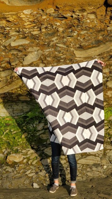 A black and white Bracken quilt is held by the maker in front of a grassy hill.