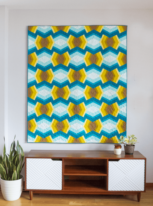 A bright teal and yellow Bracken quilt hangs on a gray wall above a brown and white lowboy.