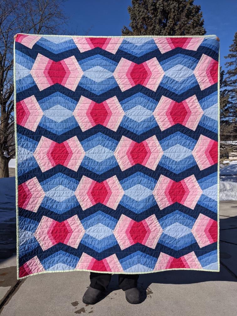 A pink and blue Bracken quilt is held by the maker outside on pavement.