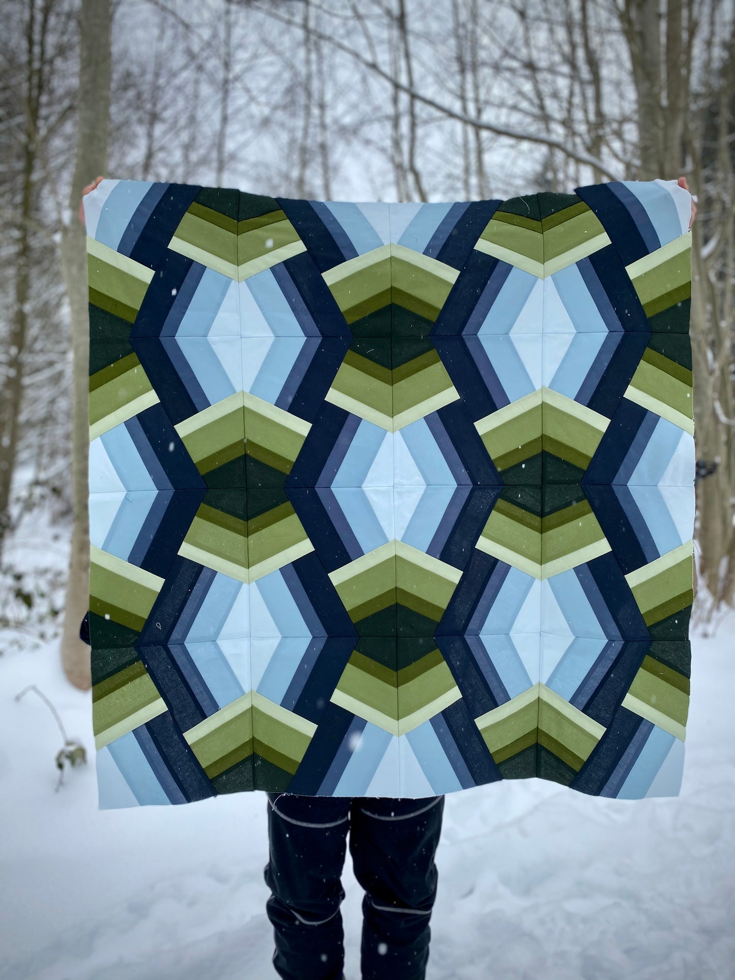 A green and blue Bracken quilt is held by the maker in the snow.